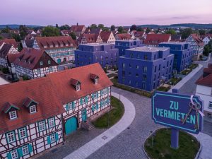 Aerial view of German residential neighborhood with red-tiled roofs, cobblestone streets, and For Rent sign in foreground