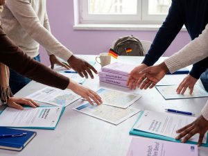 Diverse Hände arbeiten gemeinsam an einem Tisch mit deutschen Dokumenten und Willkommensmaterialien in einem einladenden Büro